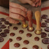 Hands using wooden chocolate stamps to press custom logo designs onto circular chocolate pieces arranged on a tray.